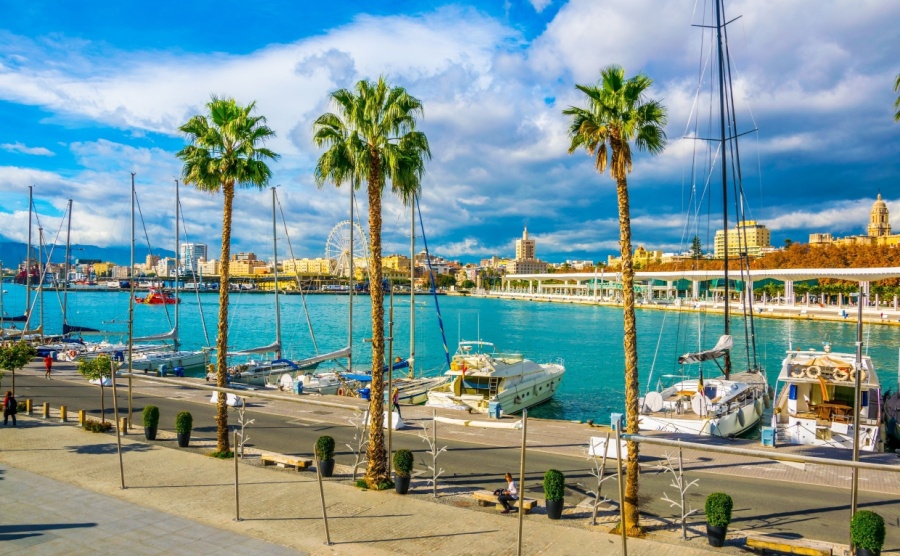 people are walking on a promenade surrounded by marina in the port of malaga in spain.