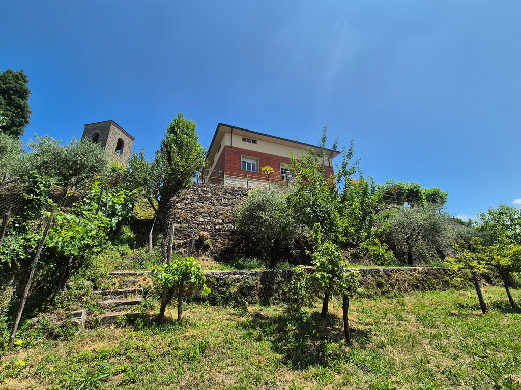 Red-rendered house above a terraced garden in Garfagnana, Tuscany, with olive trees and a stone bell tower nearby.