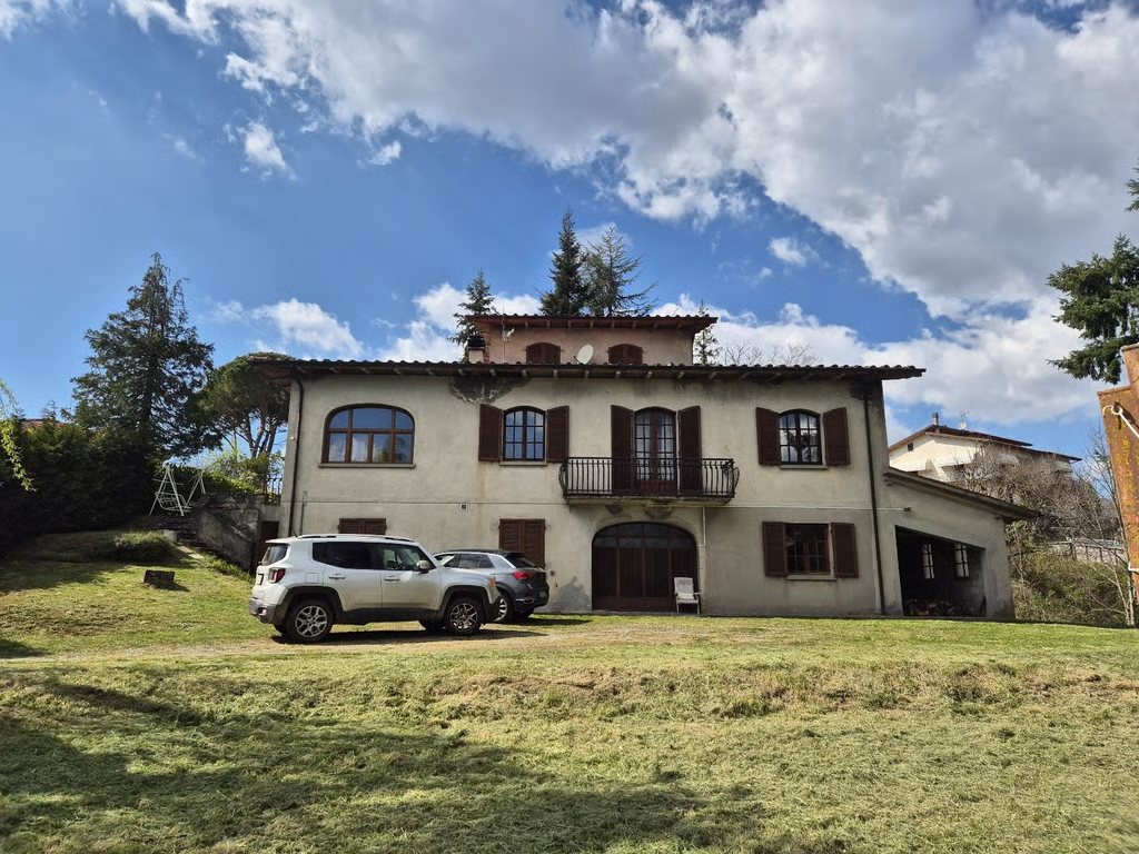 Two-storey house in Piazza al Serchio, Tuscany, with cream walls, wooden shutters, a balcony and a grassy driveway.