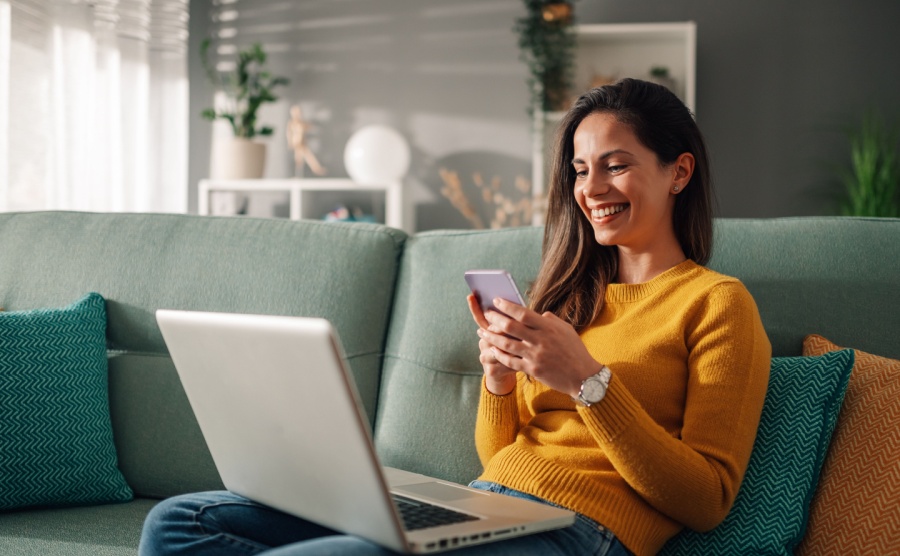 Portrait of a woman using laptop and a phone and checking email or news online while sitting on sofa at home
