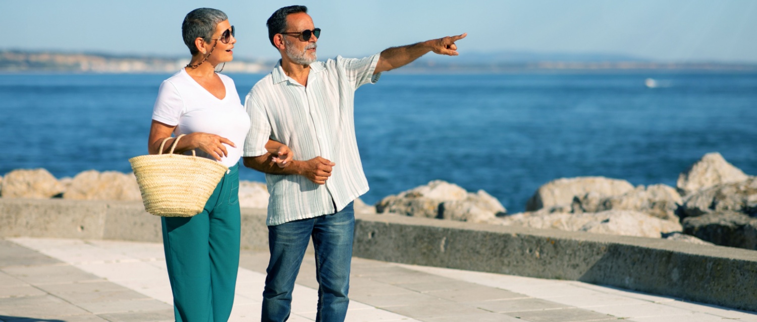 Retirement Travel. Mature Tourists Couple Pointing Finger Aside Looking At Sea View Outdoors, Walking Along Coastline And Enjoying Dream Vacation And Active Retired Life In Summer.