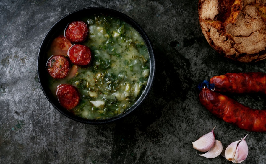 Caldo Verde Soup with greens and chopped chorizo on the top in the bowl on a dark background. Traditional round bread, chorizo and garlic.