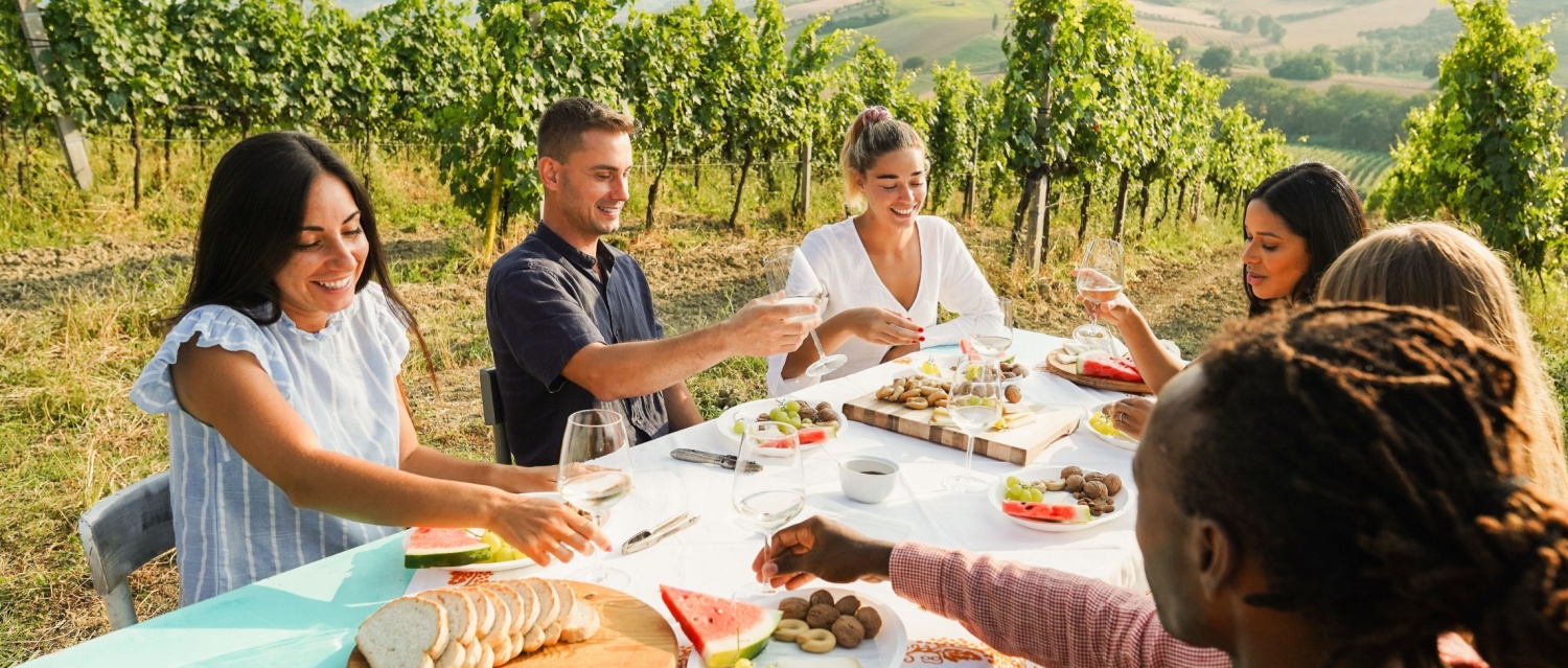 Happy adult friends having fun drinking white wine and eating together with vineyard in background - Multiracial people doing dinne at summer time in countryside resort - Main focus on blond girl face