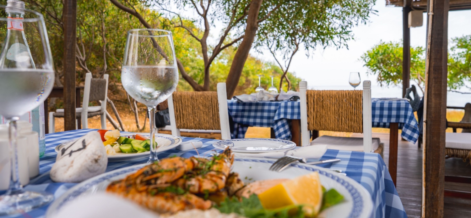 A plate of grilled seafood with lemon and greens on a blue and white tablecloth, surrounded by trees, rustic decor, and a glimpse of the sea in Cyprus.