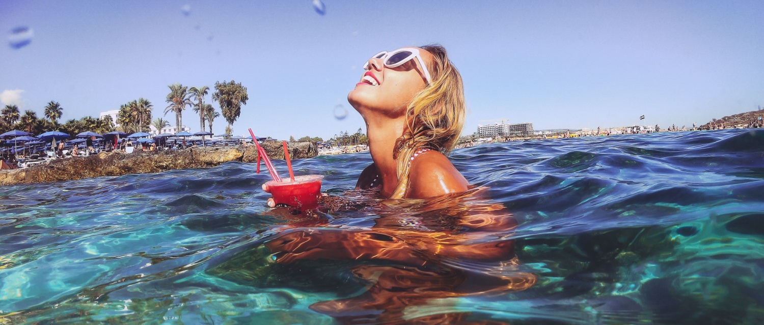 Summer lifestyle portrait of young stunning blonde girl. Enjoying life, smiling, drinking cocktail in the turquoise sea. Wearing stylish sunglasses, stripes swimsuit. Half over half under water.