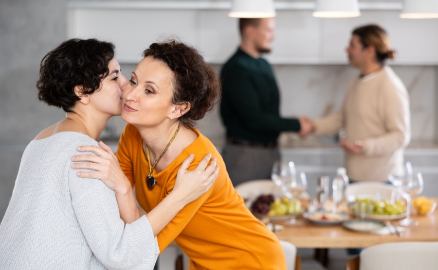 Women hug each other and kiss on the cheek while greeting each other before a festive dinner. Men shaking hands in the background