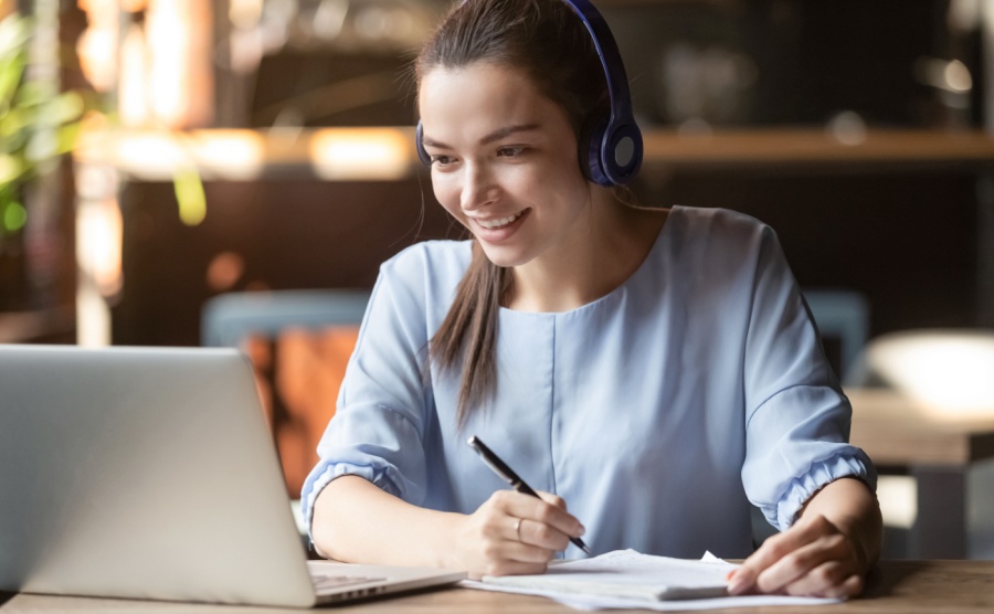 Smiling girl student wear wireless headphone study online with skype teacher, happy young woman learn language listen lecture watch webinar write notes look at laptop sit in cafe, distant education
