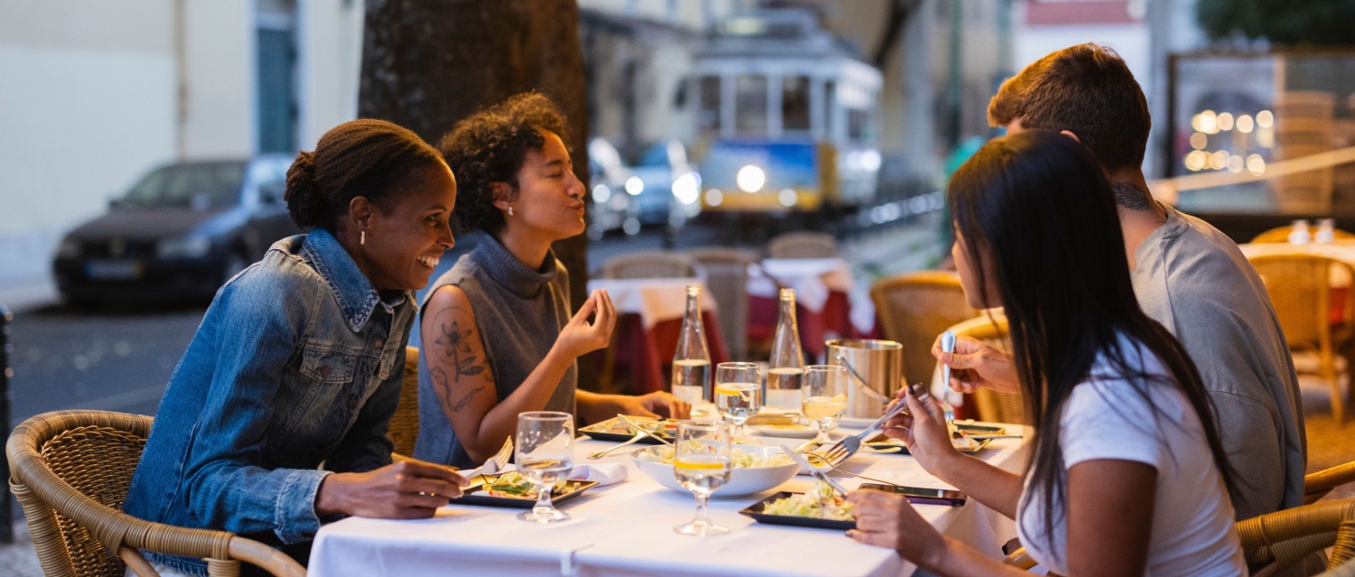 Four friends are happily eating dinner together at an outdoor restaurant in lisbon, portugal, with a traditional yellow tram passing by in the background, creating a lively urban atmosphere