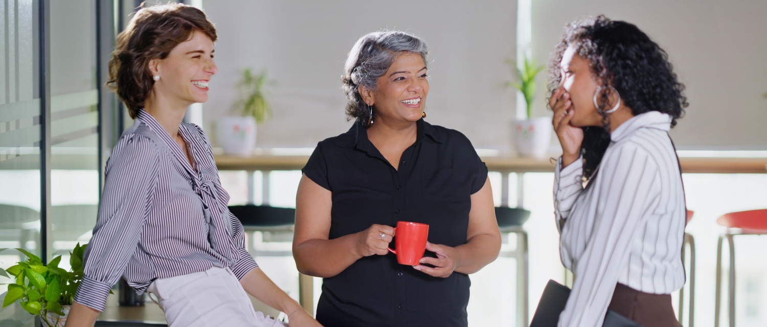 Indian smiling old business women standing hold cup drink coffee talk gossip indoor work place. Beautiful young adult corporate girls make funny blab share ideas during tea break spend time job office
