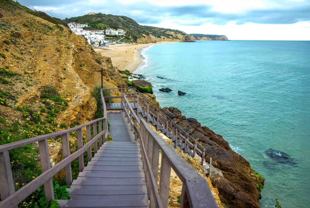 Beautiful ocean beach scene with lon wooden staircase steps that lead down the rocky cliffs to the ocean with idyllic beach and village background travel tourism Salema Beach, Algarve Portugal
