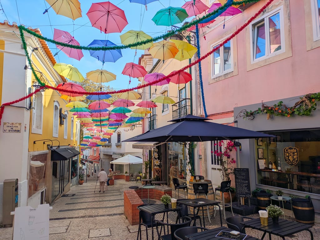 Colorful umbrellas hang above a narrow street with cafe tables, wine bar, and pastel buildings in Caldas da Rainha, Portugal