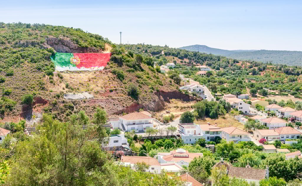 A sunny, wide view of an inland Algarve village nestled among green, tree-covered hills. In the background, a large, vibrant Portuguese flag is painted directly onto an exposed rocky hillside above traditional whitewashed houses with terracotta roofs