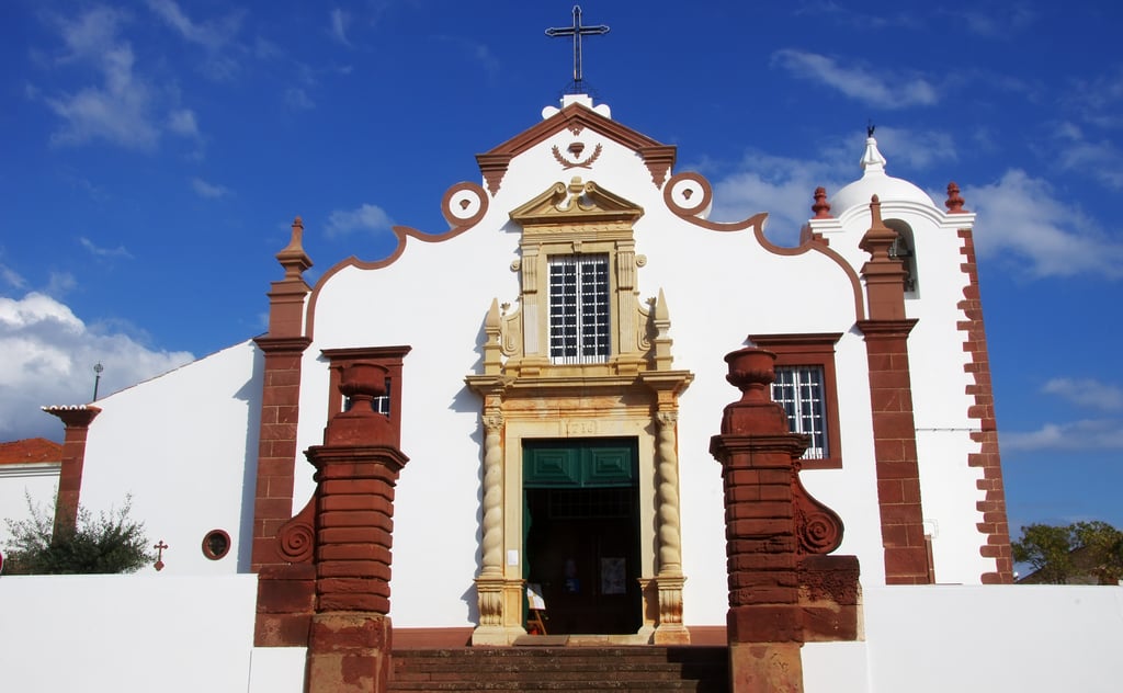 A traditional Portuguese church with a bright whitewashed facade, striking reddish-brown stone accents along its edges and pillars, an ornate entryway with green doors, and a small bell tower against a clear blue sky.