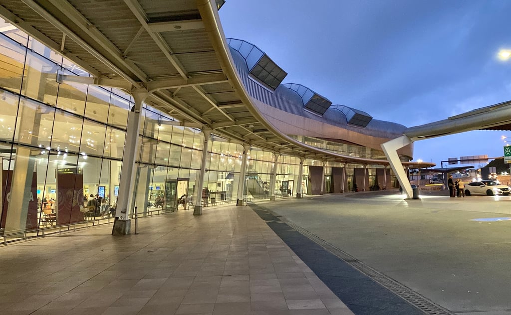 The exterior of Faro Airport at dusk, featuring a modern terminal with a sweeping curved roof structure, large glass windows revealing the softly lit interior, and a spacious, empty paved walkway in the foreground