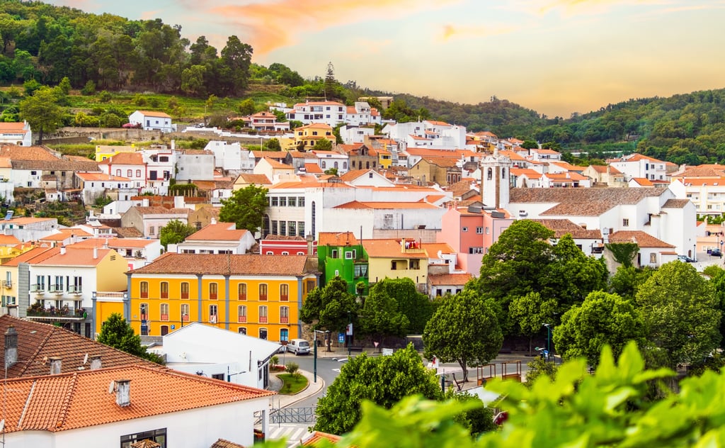 A beautiful view of the mountainous town of Monchique in the Algarve, Portugal. The town features traditional whitewashed buildings with terracotta roofs, alongside striking bright yellow, green, and pink houses, all set against a warm sunset sky and lush green forests.