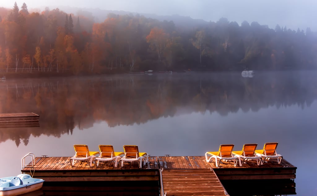 Wooden dock with lounge chairs overlooking a calm lake surrounded by forested shoreline with autumn-coloured trees, showing the serene rural setting typical of Canadian recreational properties.