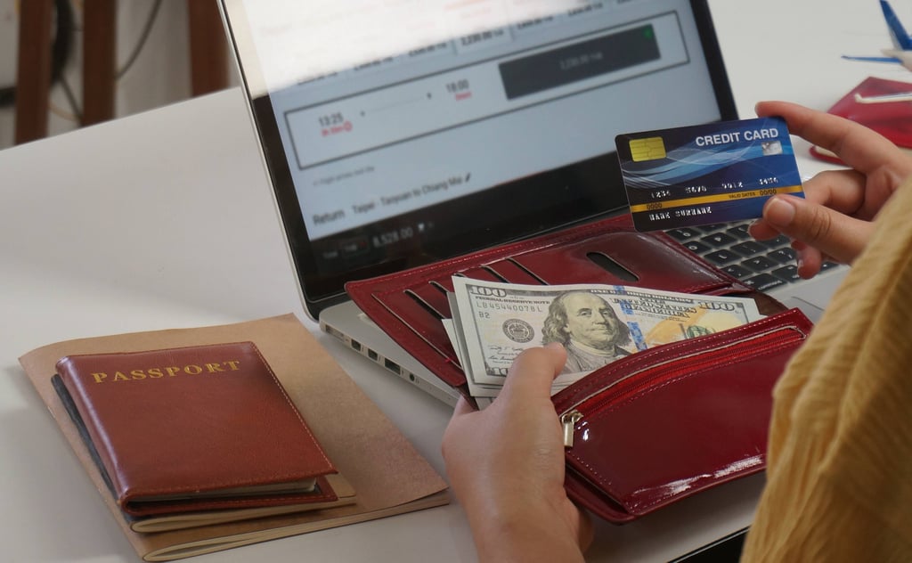 Hands holding a wallet of banknotes and a bank card at a laptop showing a travel booking