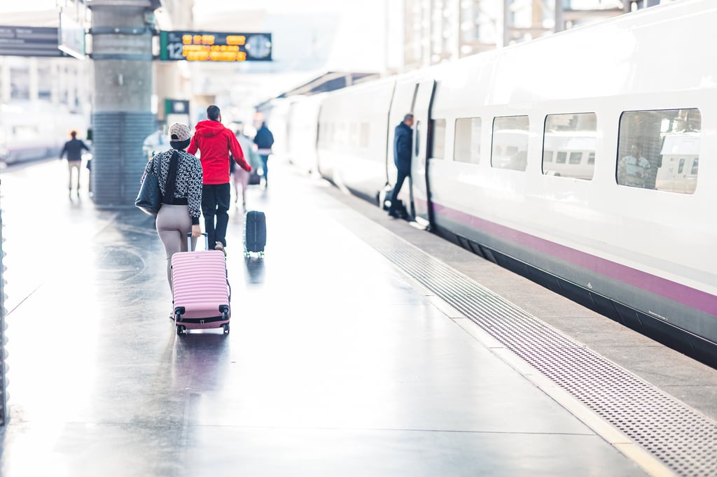 Passengers with wheeled suitcases walk along a platform beside a high-speed train in Spain