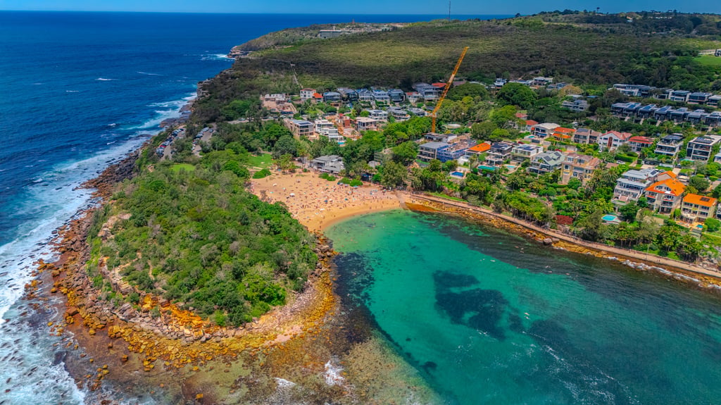 Aerial view of a sandy cove with turquoise water, a wooded headland and houses with pools beside the beach in Sydney
