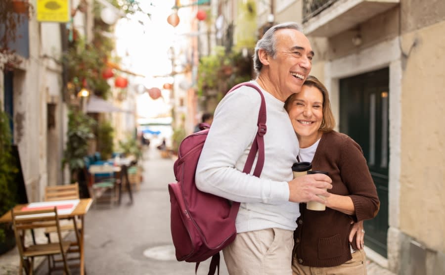 Joyful Senior Tourists Couple Hugging Posing With Backpack Holding Paper Coffee Cups On Cozy Lisbon Street With Outdoor Cafes. Retired Elderly Spouses Enjoying Vacation And Retirement In Europe