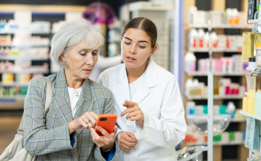 Female pharmacist and elderly woman customer reading prescription together on smartphone screen in pharmacy